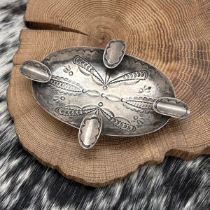 May include: Oval silver ashtray with decorative floral and geometric patterns. The ashtray has four rounded, petal-shaped rests and a central design. The metal has a slightly tarnished appearance, giving it a vintage look. The ashtray is placed on a wooden surface.