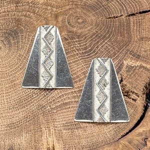 May include: Two silver-toned, trapezoid-shaped earrings with a central zigzag design and decorative accents. The earrings have a dark gray border and are displayed on a wooden surface.