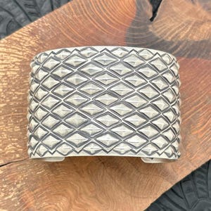May include: A wide, silver cuff bracelet featuring a repeating diamond pattern. The bracelet has a textured surface and a slightly curved shape. The cuff is displayed on a wooden surface.