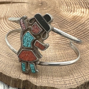 May include: A silver bracelet with a turquoise and red stone inlay depicting a stylized figure wearing a black hat.