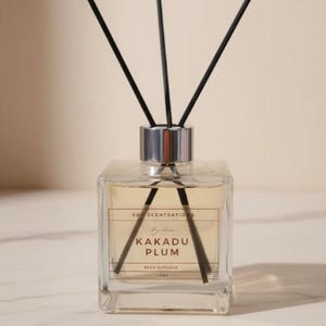 May include: A square glass reed diffuser bottle filled with light amber liquid, featuring a silver cap and three black reeds. The label reads Soy Scentsations, By Lisa, Kakadu Plum, Reed Diffuser, 150g. The bottle has a clear, minimalist design.