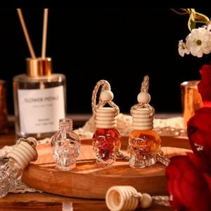 May include: Three glass skull-shaped car air fresheners with wooden tops and brown liquid inside. The air fresheners are hanging from a string with a wooden bead. The air fresheners are on a wooden surface with red roses in the background.