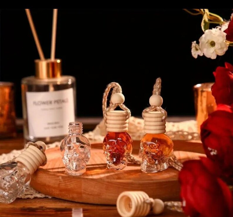 May include: Three glass skull-shaped car air fresheners with wooden tops and brown liquid inside. The air fresheners are hanging from a string with a wooden bead. The air fresheners are on a wooden surface with red roses in the background.