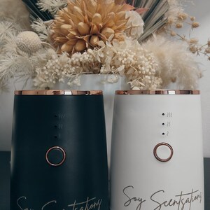 May include: Two contemporary aromatherapy diffusers, one black and one white, with gold trim. Each has a power button and the text "Soy Scentations". A dried floral arrangement is in the background.
