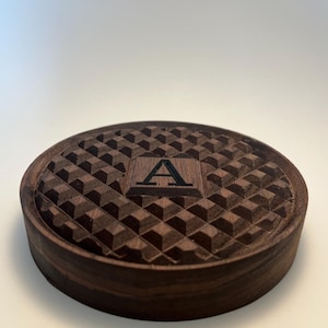 May include: A round, dark brown wooden coaster with a geometric pattern carved into the surface. The center features a square with the letter "A" engraved. The coaster is made of a dark wood and has a smooth, rounded edge.