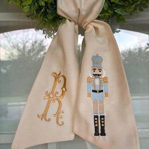 Puede incluir: Bufanda decorativa color crema con una letra "B" dorada bordada y un detallado soldado cascanueces. La bufanda está atada a una corona verde, creando una decoración festiva. El cascanueces es azul, dorado y negro.