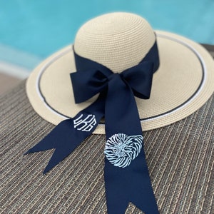 May include: A wide-brimmed straw hat with a navy blue ribbon tied in a bow. The ribbon has white embroidery of a seashell and initials.