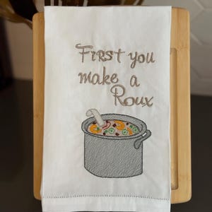 Könnte beinhalten: Weißes Küchentuch mit gesticktem Text "First you make a Roux" über einem grauen Suppentopf. Der Topf hat eine Kelle und ist mit Gemüse gefüllt. Das Handtuch wird auf einem Holzschneidebrett präsentiert.