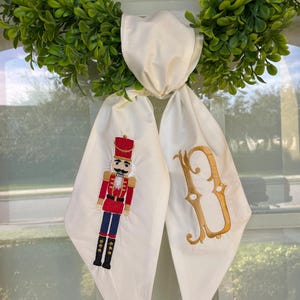 Puede incluir: Un lazo de corona decorativo blanco con una base de corona de hojas verdes. Un lado presenta un cascanueces bordado en rojo, azul y negro. El otro lado tiene una letra "B" bordada en oro.