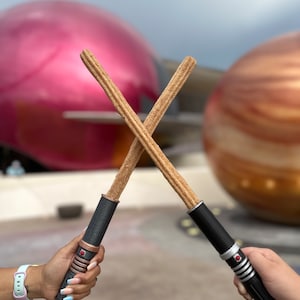 May include: Two churros are crossed like lightsabers, with black handles and red buttons. The churros are in focus, with a blurred background of a pink and brown sphere.