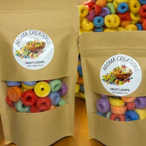 May include: Two brown paper bags with clear windows showing colorful wax melts shaped like cereal loops. The bags have labels that say "Aroma Creations" and "Fruit Loops" with the size of the wax melts in ounces.
