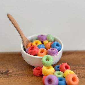 May include: A white bowl filled with colorful cereal rings, spilling out onto a wooden surface. The cereal is a variety of colors, including red, orange, yellow, green, blue, and purple.