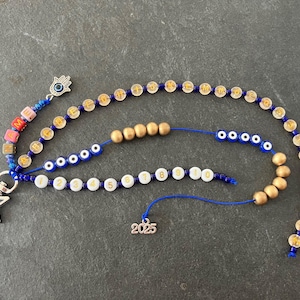 Könnte beinhalten: Eine Sammlung von Schmuckstücken mit blauen und goldenen Perlen. Der Schmuck umfasst einen Anhänger mit dem Wort "ME", einen sternförmigen Anhänger und einen Anhänger mit der Zahl "2025". Die Perlen sind in verschiedenen Mustern angeordnet.