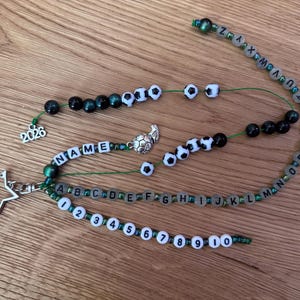 Könnte beinhalten: Ein personalisierter Schlüsselanhänger mit Perlen und einem Sternanhänger. Der Schlüsselanhänger hat Perlen mit Buchstaben, Zahlen und Fußballmotiven. Das Wort "NAME" ist auf einem Strang geschrieben, und das Jahr "2026" ist enthalten.