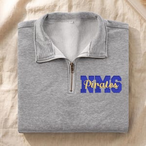 May include: A heather gray quarter-zip sweatshirt featuring a fold-over collar and a metal zipper. On the chest, the letters NMS are embroidered in blue with a gold outline, layered with the word Pirates in gold cursive script embroidery.