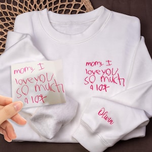 May include: White sweatshirt with the handwritten text "momy I love you so much" in pink on the front. The sleeve has the name "Olivia" embroidered in pink. A handwritten note with the same text is held in front.