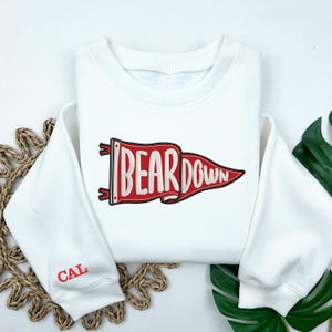May include: White sweatshirt with a red pennant design that reads "BEAR DOWN." The left sleeve has "CAL" embroidered in red. The sweatshirt is folded on a woven mat with green leaves in the background.