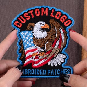 May include: Embroidered patch featuring a bald eagle with outstretched wings, an American flag, and the text "CUSTOM LOGO" in red. The patch has a blue border and the words "EMBROIDED PATCHES" at the bottom.