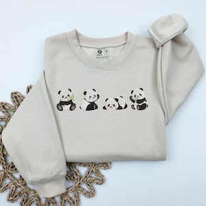 May include: A beige sweatshirt with an embroidered design of four pandas holding bamboo shoots. The pandas are black and white with green bamboo shoots.