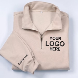 May include: A beige pullover sweatshirt with a quarter-zip neckline. The front features the text "YOUR LOGO HERE" in black. The left sleeve cuff displays the words "Custom text" in black. The sweatshirt is folded neatly.
