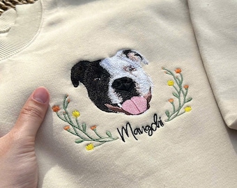 May include: Cream-colored sweatshirt with an embroidered dog portrait. The dog has black and white fur, a pink tongue, and is surrounded by a floral wreath. The name "Mangchi" is embroidered below the dog.