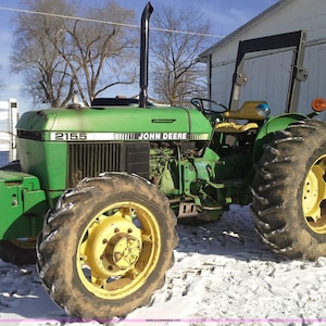 May include: A green John Deere 2155 tractor with black exhaust pipe and yellow wheels. The tractor is parked in a snowy field. The tractor has the words "John Deere" printed on the side.
