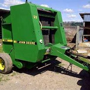 May include: A green John Deere round baler with a yellow stripe and a black hitch. The baler has a large, rectangular bale chamber and a series of rollers that compress the hay into a bale.