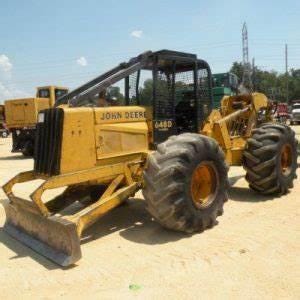 Pode incluir: Uma máquina florestal John Deere 648D amarela com uma cabine preta e uma grande lâmina na frente. A máquina tem pneus grandes e está sentada em uma superfície de terra.