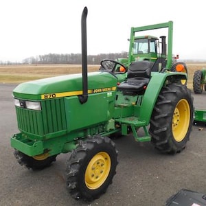 May include: A green John Deere 870 tractor with a black exhaust pipe. The tractor has a black seat and a yellow steering wheel. The tractor is on a paved surface.