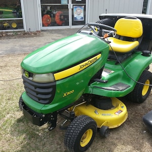 May include: A green John Deere X300 riding lawnmower with a yellow seat and black grass catcher. The mower is on a grassy surface.