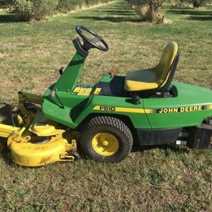 May include: A green John Deere riding lawnmower with a yellow deck and a black seat. The mower has a large rear tire and a smaller front tire. The mower has a black steering wheel and a black handle.