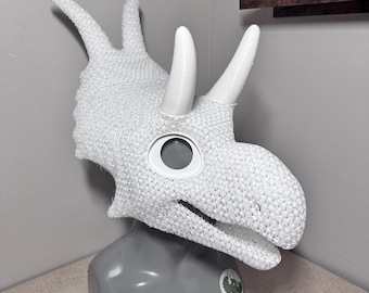 Könnte beinhalten: Eine weiße Triceratops-Dinosaurier-Maske mit drei Hörnern und einer großen Augenöffnung. Die Maske besteht aus einem strukturierten, gewebten Material und wird auf einem grauen Schaufensterkopf präsentiert. Die Hörner sind glatt und weiß.