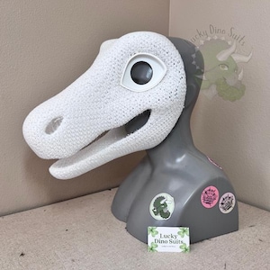 May include: A white textured dinosaur mask featuring a smooth white eye socket and open mouth, displayed on a gray mannequin head. The mannequin is decorated with several circular stickers and a small rectangular sign reading Lucky Dino Suits.