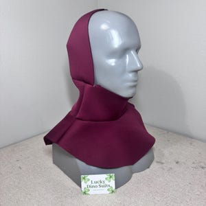 May include: A solid deep burgundy hood and neck cowl covering the head and shoulders of a grey mannequin. The fabric has a smooth, matte finish. A small white rectangular card with green clover graphics and the text Lucky Dino Suits is visible.