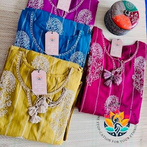 May include: Four different coloured cotton tunics with white floral embroidery and tassel detailing. The tunics are folded and arranged on a textured surface. The text "One Stop Destination for Your Ethnic" is visible in the bottom right corner of the image.