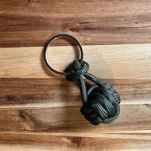Puede incluir: Un llavero de paracord verde oscuro con un anillo de llave plateado. El paracord está atado en un nudo, creando una pequeña forma de bola.