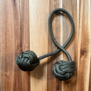 May include: A dark green decorative knot, likely made of paracord, with two spherical knots connected by a looped cord. The knots are tightly woven, and the cord has a textured appearance. The background is a wooden surface.