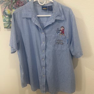 Könnte beinhalten: Hellblaues Button-Down-Hemd mit kleinem weißem Karomuster. Das Hemd hat eine Cartoon-Grafik eines Zwergs mit dem Text "Do I look like PEOPLE PERSON?" auf der Tasche. Das Hemd hängt an einem weißen Kleiderbügel.