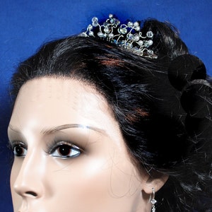 Puede incluir: Una tiara plateada con detalles de cristal y perlas se coloca sobre la cabeza de un maniqu&iacute;. El maniqu&iacute; tiene el pelo oscuro recogido en un mo&ntilde;o. El maniqu&iacute; lleva un par de pendientes plateados con una sola perla.