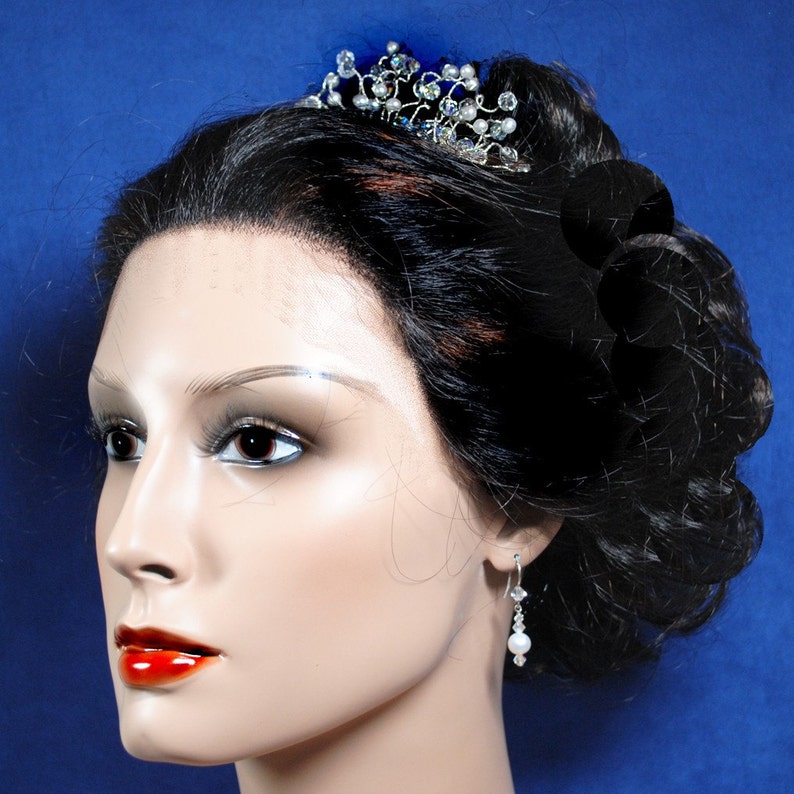 Puede incluir: Una tiara plateada con detalles de cristal y perlas se coloca sobre la cabeza de un maniqu&iacute;. El maniqu&iacute; tiene el pelo oscuro recogido en un mo&ntilde;o. El maniqu&iacute; lleva un par de pendientes plateados con una sola perla.