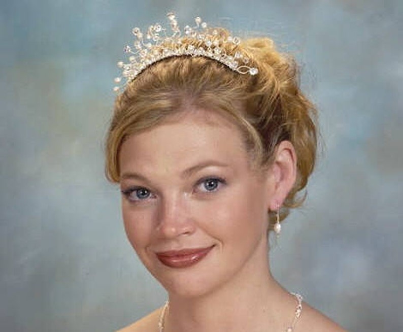 Puede incluir: Una mujer con una tiara plateada con detalles de cristal y pendientes de perla. Su cabello est&aacute; recogido en un mo&ntilde;o.