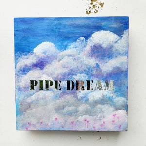 May include: A blue and white painted wooden sign with the words "PIPE DREAM" in black lettering. The background is a sky with clouds and pink flowers.