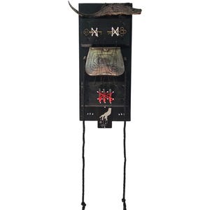 May include: A vertical black wooden wall hanging featuring a small alligator figure at the top, two brass keys tied with white thread, a metallic purse, three black cylinders wrapped in red thread, and a hand illustration with hanging tassels.