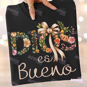 May include: Black sweatshirt featuring the phrase "Dios es Bueno" in a cursive font. The design includes floral wreaths and a bow with ribbons in shades of peach, orange, and pink. The sweatshirt has a crew neck.
