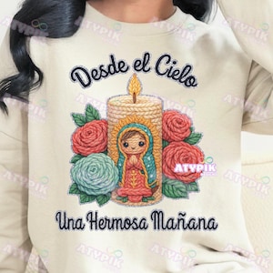 May include: Cream-colored sweatshirt featuring a graphic of a candle with a religious figure, surrounded by red and teal roses. The text "Desde el Cielo" and "Una Hermosa Mañana" are arched above and below the graphic.