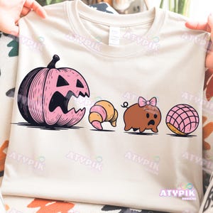 May include: Beige t-shirt featuring a pink pumpkin, croissant, pig with a bow, and a concha. The graphic design has black outlines. The text "ATYPIK DESIGNS" is printed at the bottom of the shirt.