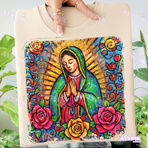 May include: Beige sweatshirt featuring a vibrant, colorful illustration of a religious figure with hands clasped in prayer. The artwork is framed by a border of roses and floral designs in shades of red, pink, yellow, and blue.