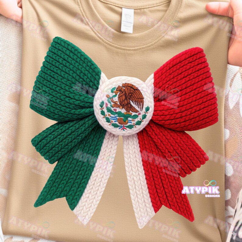 May include: A tan t-shirt with a large knitted bow in the colours of the Mexican flag: green, white, and red. The bow has a central circular emblem with the Mexican coat of arms. The design is centered on the shirt.