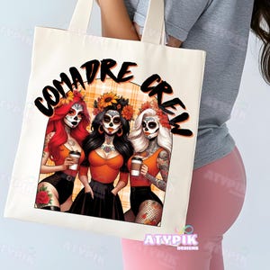 May include: A white tote bag with the text "Comadre Crew" in black lettering. The bag features an illustration of three women with sugar skulls, wearing orange tops and black skirts. The background is a plaid pattern in orange and white.