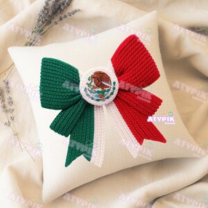 May include: A square white cushion featuring a large bow design in the colours of the Mexican flag: green, white, and red. The bow is centered and features a circular emblem with the Mexican coat of arms. The cushion is on a neutral-coloured surface.